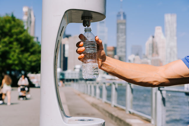 Hand filling a water bottle at a modern outdoor bottle filling station with a city skyline behind